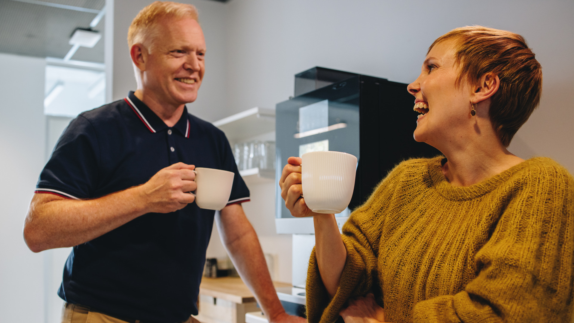En mannlig og kvinnelig kollega ler og samtaler foran kaffemaskinen på jobben